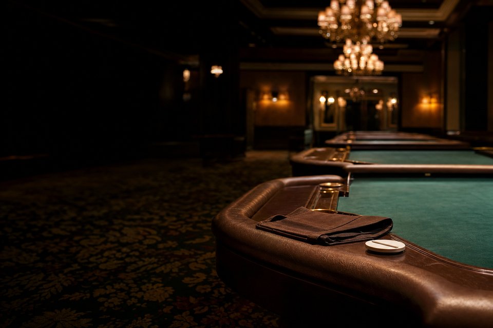 An empty craps pit at dawn, three table rails in a row under dimmed chandeliers, a folded apron and a puck on the nearest (AI-generated image)