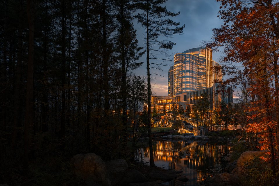 A large resort casino glowing through autumn woodland at dusk, its lit windows reflected in a still pond (AI-generated image)