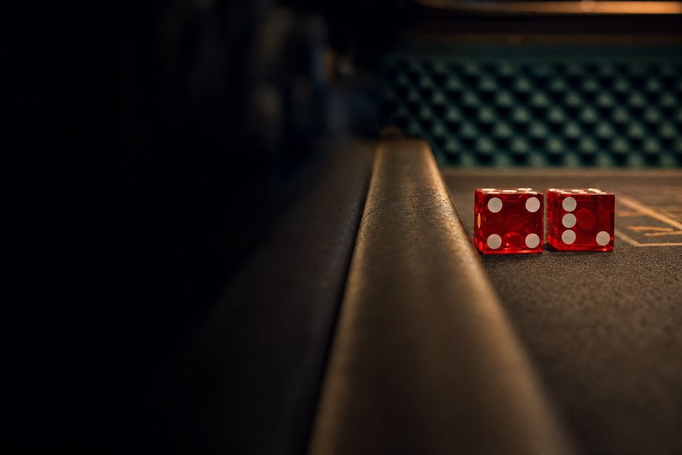 Two red casino dice held in a careful set position just above the rail of a craps table, the back wall's rubber pyramids blurred in the distance (AI-generated image)