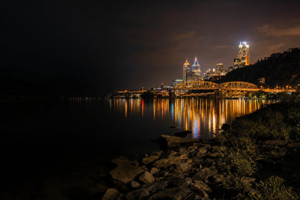 A city riverfront at night where three rivers meet, steel bridges lit gold and reflected in dark water below a hillside (AI-generated image)