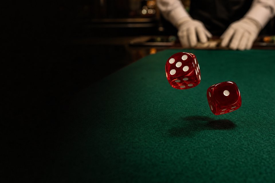 Two red dice caught mid-air above a green craps layout in a dim room, a dealer's hands blurred behind them (AI-generated image)