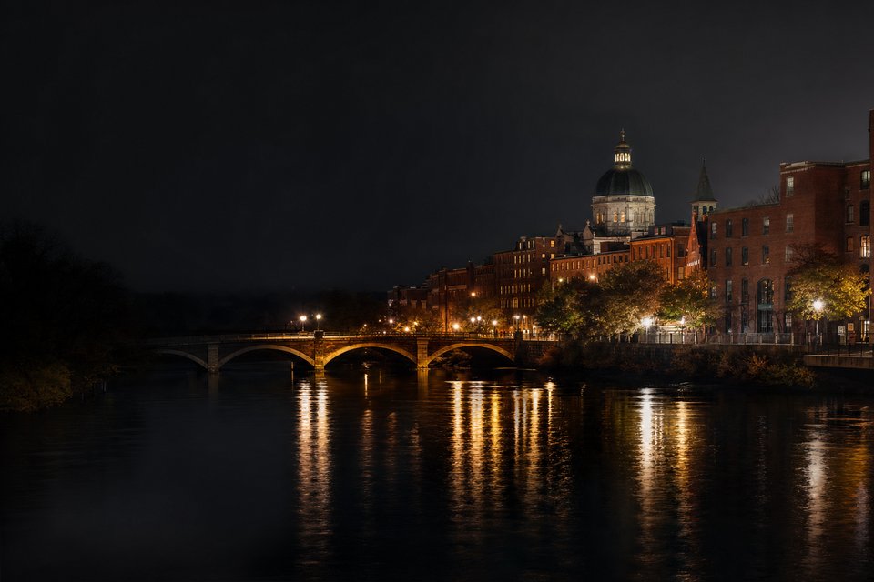 A small New England city at night seen across a calm river, old brick buildings and bridge lights reflected in the water (AI-generated image)